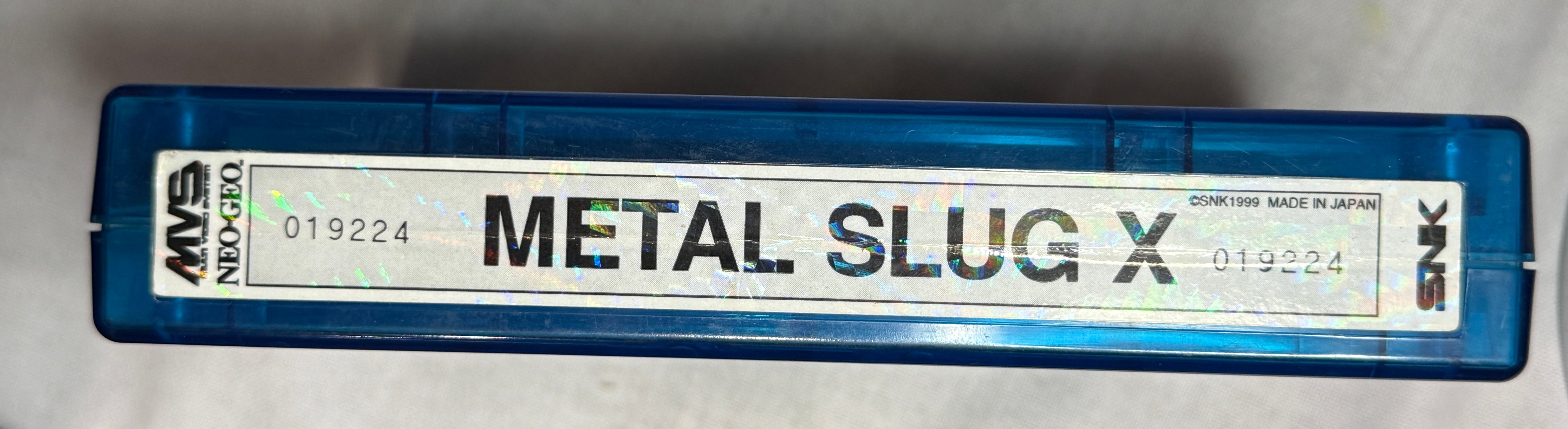 A blue translucent cassette-style case with a white label reading 'METAL SLUG X' and 'AWM', photographed on a white fabric surface.